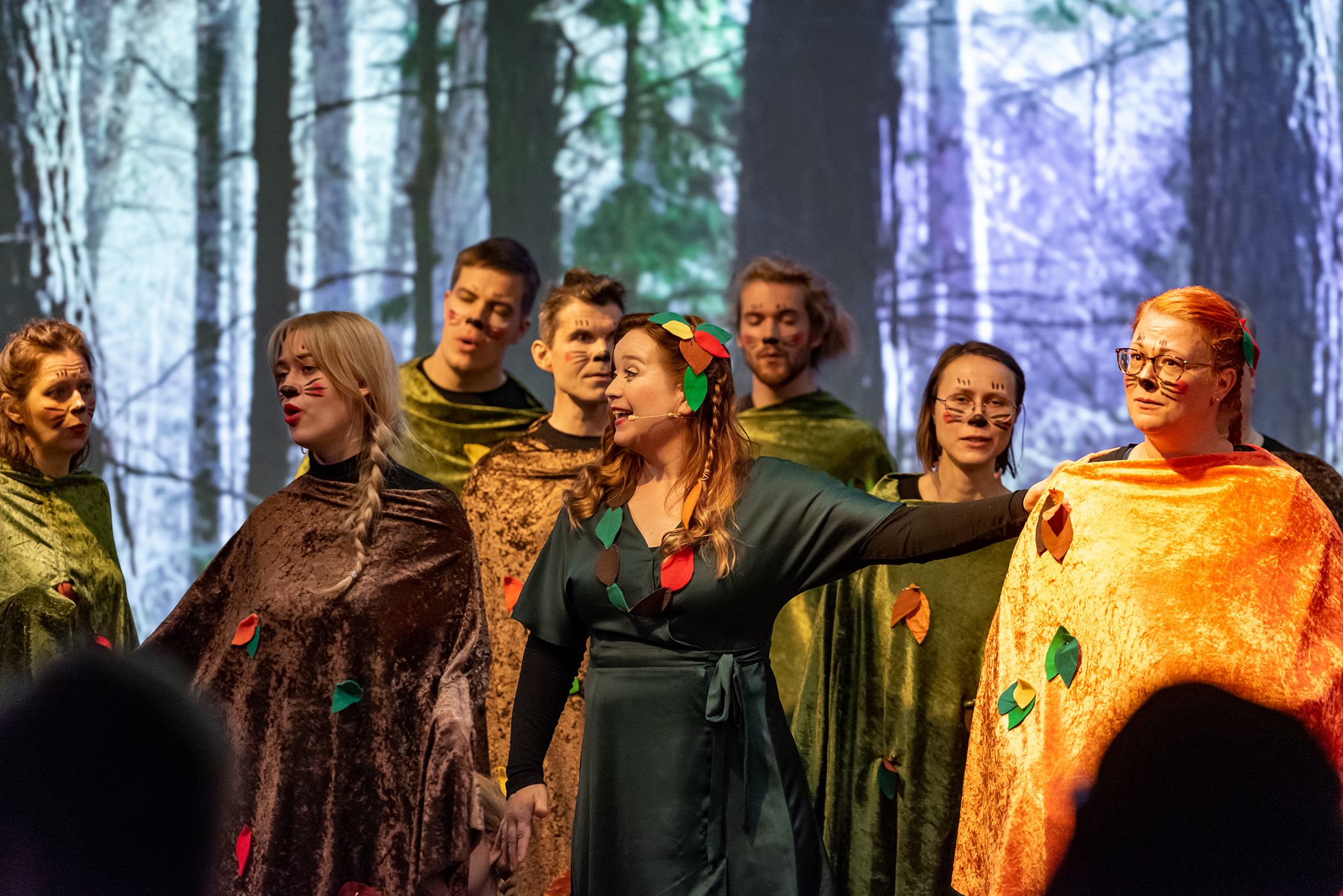 Kuorolaisia ruskeisiin ja vihreisiin kaapuihin pukeutuneena esiintyy Isa-Eerika Lehdon kanssa, taustalla iso valokuva metsästä