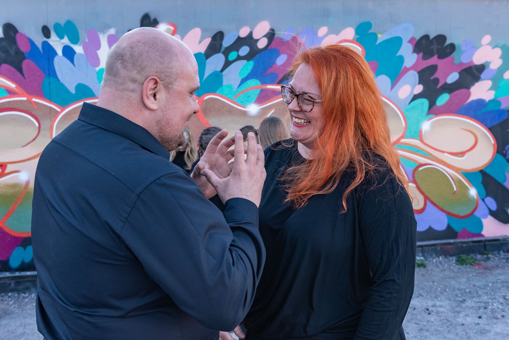 Mies ja hymyilevä, punatukkainen nainen keskustelevat graffitiseinän edessä